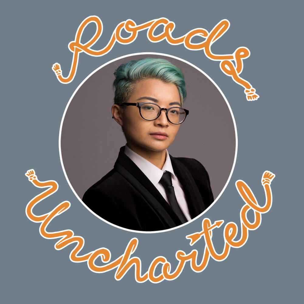 A headshot of Sebastian. They are Asian with short teal hair, wear a black suit and tie, and wear glasses. Image is framed in a white circle with orange "Roads Uncharted" surrounding it.